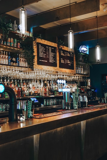 A dimly lit bar with a long counter, various bottles on shelves, hanging wine glasses, and illuminated pendant lights. The décor includes a chalkboard-style menu and beer taps glowing in blue light.