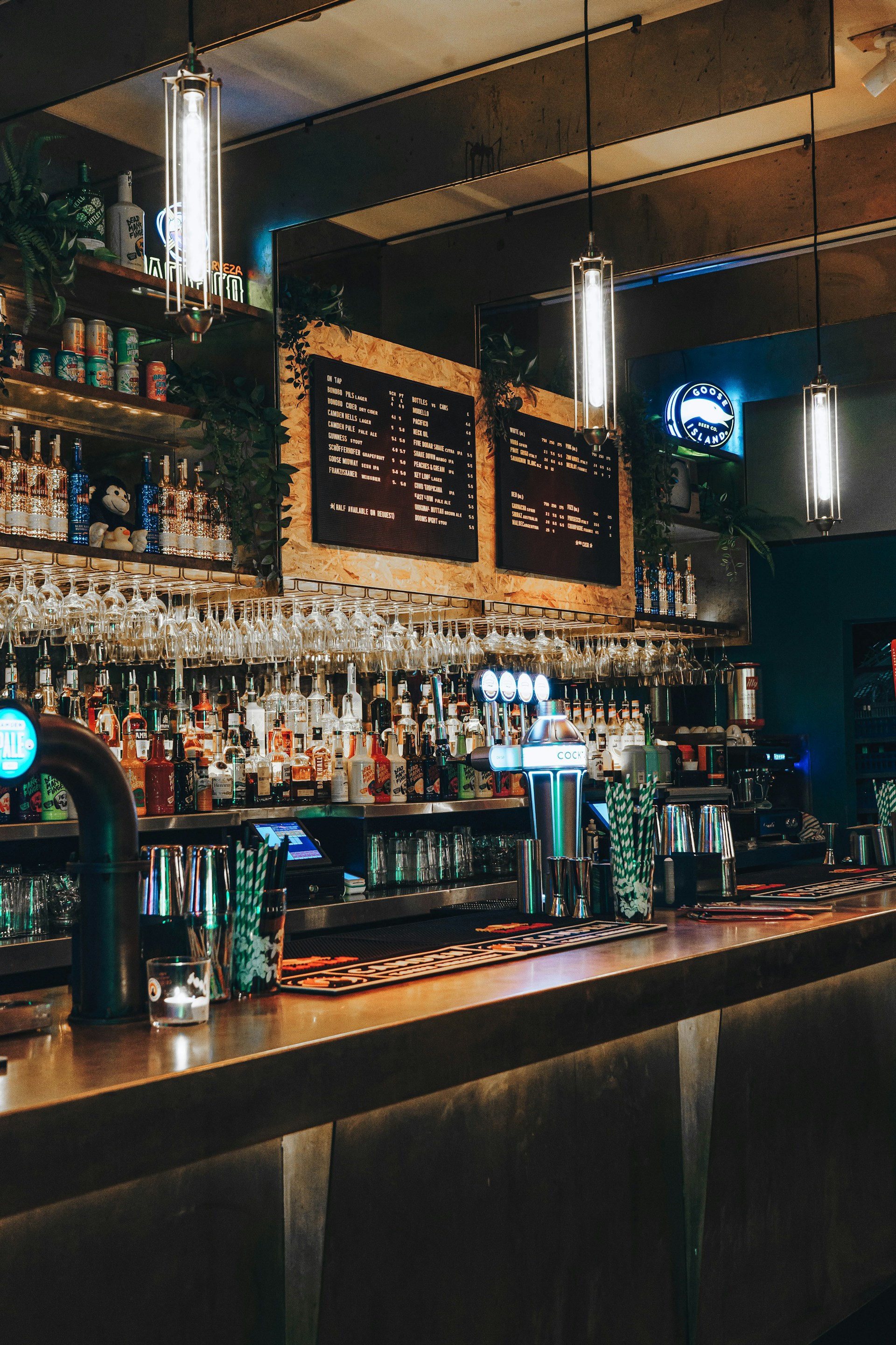A dimly lit bar with a long counter, various bottles on shelves, hanging wine glasses, and illuminated pendant lights. The décor includes a chalkboard-style menu and beer taps glowing in blue light.