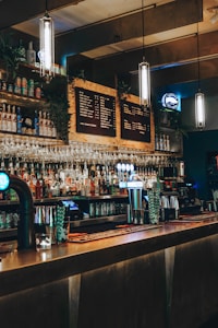 A dimly lit bar with a long counter, various bottles on shelves, hanging wine glasses, and illuminated pendant lights. The décor includes a chalkboard-style menu and beer taps glowing in blue light.