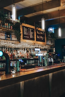 A dimly lit bar with a long counter, various bottles on shelves, hanging wine glasses, and illuminated pendant lights. The d&eacute;cor includes a chalkboard-style menu and beer taps glowing in blue light.