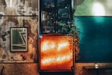 A stylish interior with a mix of industrial and cozy elements. A neon sign reading 'Wish you were here' glows warmly amidst textured walls. There is a framed abstract artwork on the wall, a modern light fixture, and lush greenery adding a touch of nature.