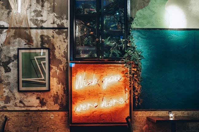 A stylish interior with a mix of industrial and cozy elements. A neon sign reading 'Wish you were here' glows warmly amidst textured walls. There is a framed abstract artwork on the wall, a modern light fixture, and lush greenery adding a touch of nature.