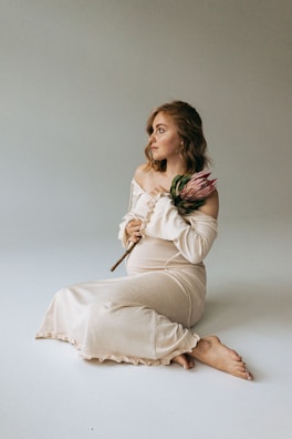 A pregnant woman is sitting on the ground with her legs crossed. She is wearing an off-shoulder, light beige dress and holding a single, large protea flower. The background is plain and neutral, creating a calm and serene setting. Her gaze is directed to the side, giving a thoughtful expression.