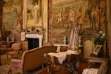 An opulent room richly adorned with elaborate tapestries and antique furnishings. The walls are decorated with intricate murals depicting historical scenes, and a large portrait in a gilded frame hangs above a classic marble fireplace. Ornate chairs, a cradle or decorative furniture piece with intricate carvings, and luxurious drapery contribute to the luxurious ambiance. Floral arrangements and decorative vases add splashes of color.