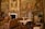 An opulent room richly adorned with elaborate tapestries and antique furnishings. The walls are decorated with intricate murals depicting historical scenes, and a large portrait in a gilded frame hangs above a classic marble fireplace. Ornate chairs, a cradle or decorative furniture piece with intricate carvings, and luxurious drapery contribute to the luxurious ambiance. Floral arrangements and decorative vases add splashes of color.