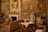 An opulent room richly adorned with elaborate tapestries and antique furnishings. The walls are decorated with intricate murals depicting historical scenes, and a large portrait in a gilded frame hangs above a classic marble fireplace. Ornate chairs, a cradle or decorative furniture piece with intricate carvings, and luxurious drapery contribute to the luxurious ambiance. Floral arrangements and decorative vases add splashes of color.