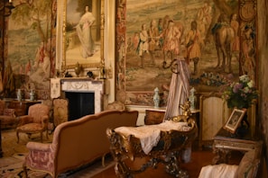 An opulent room richly adorned with elaborate tapestries and antique furnishings. The walls are decorated with intricate murals depicting historical scenes, and a large portrait in a gilded frame hangs above a classic marble fireplace. Ornate chairs, a cradle or decorative furniture piece with intricate carvings, and luxurious drapery contribute to the luxurious ambiance. Floral arrangements and decorative vases add splashes of color.