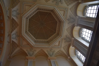 A ceiling features an ornate octagonal design with intricate patterns and detailed moldings. The architecture includes arched windows allowing natural light to enter, casting subtle shadows on the walls. The color scheme consists of neutral tones with hints of gold and beige, enhancing the classical elegance.