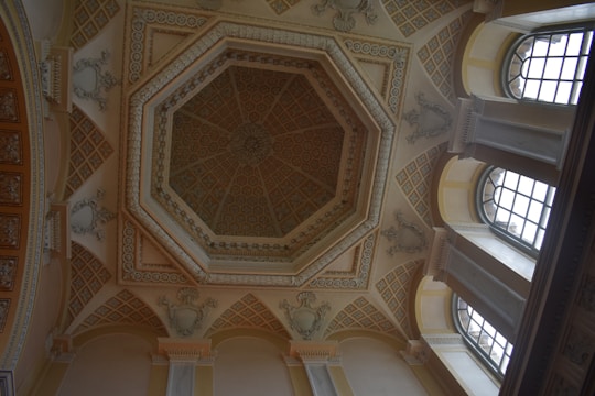 A ceiling features an ornate octagonal design with intricate patterns and detailed moldings. The architecture includes arched windows allowing natural light to enter, casting subtle shadows on the walls. The color scheme consists of neutral tones with hints of gold and beige, enhancing the classical elegance.