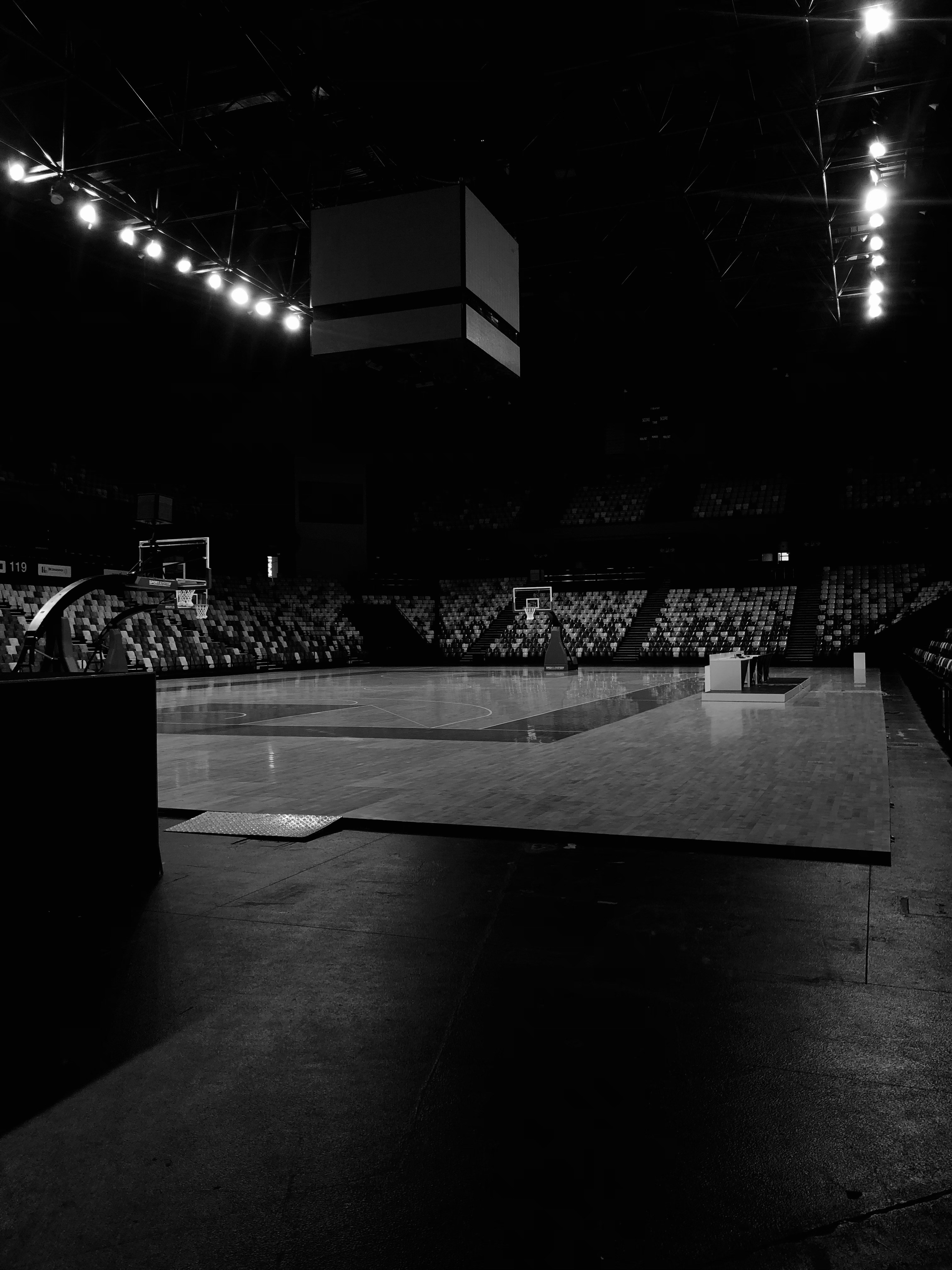 A dimly lit indoor basketball court with an empty wooden floor, flanked by seating areas with patterned designs. Overhead, there is a scoreboard suspended from the ceiling, and bright lights line the upper framework providing minimal illumination.
