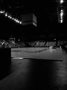A dimly lit indoor basketball court with an empty wooden floor, flanked by seating areas with patterned designs. Overhead, there is a scoreboard suspended from the ceiling, and bright lights line the upper framework providing minimal illumination.