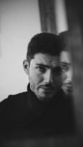 A monochrome portrait of a man with a contemplative expression, partly reflected in a mirror. The focus is on his eyes, and the composition uses lighting to create a moody, intimate atmosphere.
