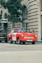A vintage red car is parked on a city street near a tall tree and a building with arched windows. The car has a sign on the side indicating it's used for sightseeing tours. Additional cars are parked nearby, and a sign for a computer store can be seen on the building.
