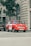 A vintage red car is parked on a city street near a tall tree and a building with arched windows. The car has a sign on the side indicating it's used for sightseeing tours. Additional cars are parked nearby, and a sign for a computer store can be seen on the building.
