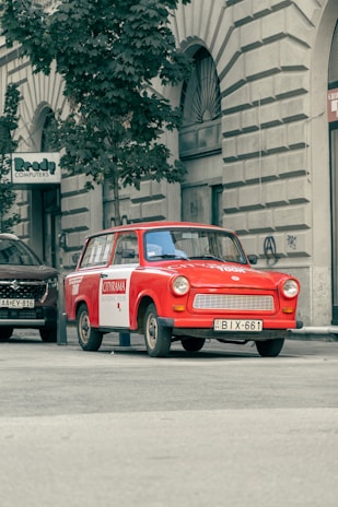 A vintage red car is parked on a city street near a tall tree and a building with arched windows. The car has a sign on the side indicating it's used for sightseeing tours. Additional cars are parked nearby, and a sign for a computer store can be seen on the building.