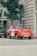 A vintage red car is parked on a city street near a tall tree and a building with arched windows. The car has a sign on the side indicating it's used for sightseeing tours. Additional cars are parked nearby, and a sign for a computer store can be seen on the building.