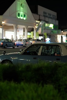 A nighttime scene featuring a blue taxi parked in front of a building with illuminated signage. Several other vehicles are visible in the background, indicating a lively area. The building has a triangular roof with green-lit elements, and there are shrubs in the foreground.