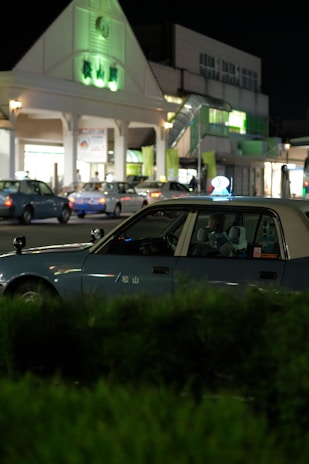 A nighttime scene featuring a blue taxi parked in front of a building with illuminated signage. Several other vehicles are visible in the background, indicating a lively area. The building has a triangular roof with green-lit elements, and there are shrubs in the foreground.