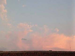 A tile roof is silhouetted against a sky filled with soft pink and blue clouds. The sky is light and calm, suggesting a time around sunset or sunrise. An antenna is visible on the right side, slightly above the roofline.