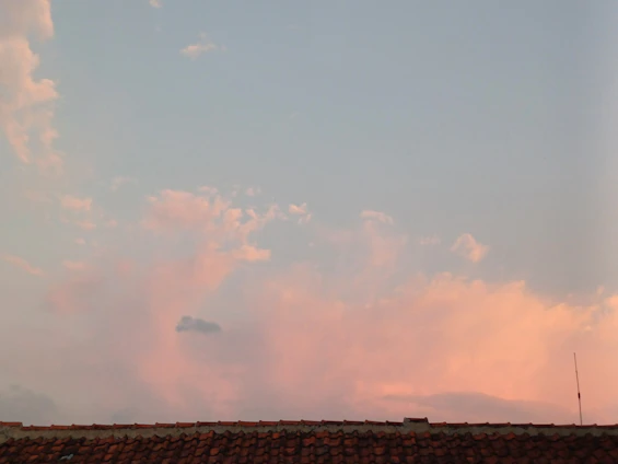 A tile roof is silhouetted against a sky filled with soft pink and blue clouds. The sky is light and calm, suggesting a time around sunset or sunrise. An antenna is visible on the right side, slightly above the roofline.