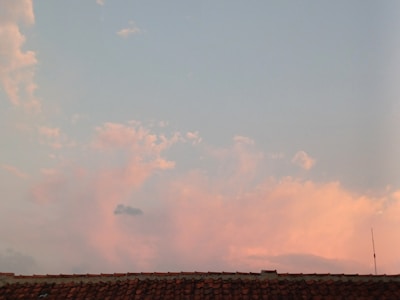 A tile roof is silhouetted against a sky filled with soft pink and blue clouds. The sky is light and calm, suggesting a time around sunset or sunrise. An antenna is visible on the right side, slightly above the roofline.