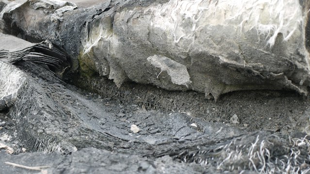 A close-up view of a worn and eroded surface with layers of what appears to be asbestos fibers and crumbling material. The texture is rough, with a mix of dark and light gray shades, and some white fibers protruding from the surface.