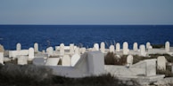 A serene coastal cemetery with rows of white tombstones set against the backdrop of a vast blue sea. The tombstones are unevenly spaced among patches of grass and wild plants, creating a peaceful and contemplative atmosphere.