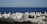 A serene coastal cemetery with rows of white tombstones set against the backdrop of a vast blue sea. The tombstones are unevenly spaced among patches of grass and wild plants, creating a peaceful and contemplative atmosphere.