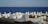A serene coastal cemetery with rows of white tombstones set against the backdrop of a vast blue sea. The tombstones are unevenly spaced among patches of grass and wild plants, creating a peaceful and contemplative atmosphere.