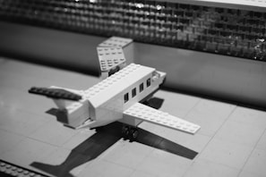 A monochrome image features a small model airplane constructed from LEGO bricks. The plane is positioned on a flat surface with a tiled pattern, and in the background, a textured wall is visible.