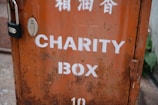 A weathered metal charity box with an orange hue and white lettering. The box is secured with a black padlock and shows signs of rust and wear. Above the English text, there are markings in another language, possibly Chinese.