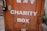 A weathered metal charity box with an orange hue and white lettering. The box is secured with a black padlock and shows signs of rust and wear. Above the English text, there are markings in another language, possibly Chinese.
