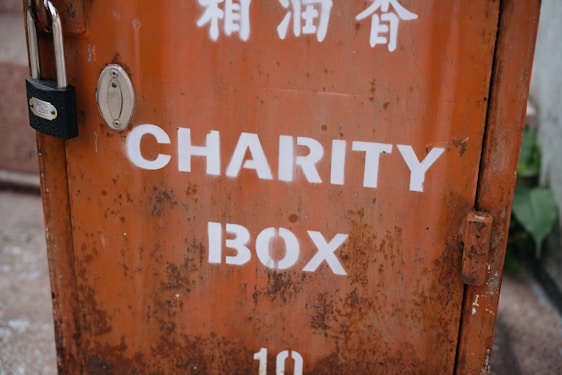 A weathered metal charity box with an orange hue and white lettering. The box is secured with a black padlock and shows signs of rust and wear. Above the English text, there are markings in another language, possibly Chinese.
