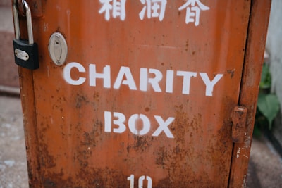 A weathered metal charity box with an orange hue and white lettering. The box is secured with a black padlock and shows signs of rust and wear. Above the English text, there are markings in another language, possibly Chinese.
