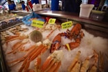 A display of fresh seafood on ice, including crab legs, a giant wild lobster, and a sea urchin. The seafood is labeled with bright yellow signs, and a Norwegian flag is visible in the background. People in blue uniforms are seen behind the counter.