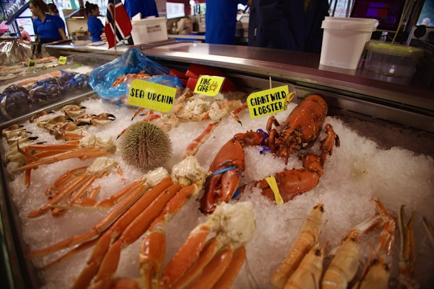 A display of fresh seafood on ice, including crab legs, a giant wild lobster, and a sea urchin. The seafood is labeled with bright yellow signs, and a Norwegian flag is visible in the background. People in blue uniforms are seen behind the counter.