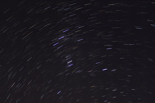 A long-exposure photograph of the night sky, capturing star trails forming circular patterns. The background is a deep black, accentuating the luminous streaks of stars in white, blue, and faint yellow hues.