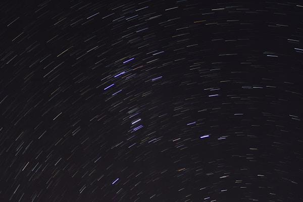 A long-exposure photograph of the night sky, capturing star trails forming circular patterns. The background is a deep black, accentuating the luminous streaks of stars in white, blue, and faint yellow hues.