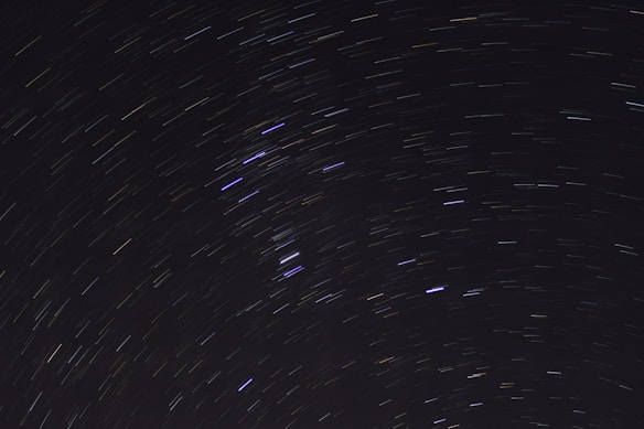 A long-exposure photograph of the night sky, capturing star trails forming circular patterns. The background is a deep black, accentuating the luminous streaks of stars in white, blue, and faint yellow hues.