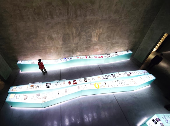 An indoor exhibition space features zigzag-shaped display cases with a variety of small artifacts and items, illuminated with bright lighting. The setting has a modern and minimalistic aesthetic with grey concrete walls and floor. A person is observing the displays closely, while another figure can be seen walking in the background.