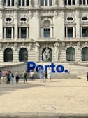 A large, ornate building with intricate architectural details and multiple arched windows serves as the backdrop. In front of the building, there is a prominent blue sign spelling 'Porto'. Several people are walking and standing around the area, creating a lively atmosphere.