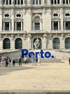 A large, ornate building with intricate architectural details and multiple arched windows serves as the backdrop. In front of the building, there is a prominent blue sign spelling 'Porto'. Several people are walking and standing around the area, creating a lively atmosphere.