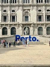 A large, ornate building with intricate architectural details and multiple arched windows serves as the backdrop. In front of the building, there is a prominent blue sign spelling 'Porto'. Several people are walking and standing around the area, creating a lively atmosphere.