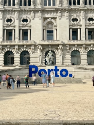 A large, ornate building with intricate architectural details and multiple arched windows serves as the backdrop. In front of the building, there is a prominent blue sign spelling 'Porto'. Several people are walking and standing around the area, creating a lively atmosphere.