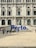 A large, ornate building with intricate architectural details and multiple arched windows serves as the backdrop. In front of the building, there is a prominent blue sign spelling 'Porto'. Several people are walking and standing around the area, creating a lively atmosphere.