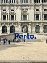 A large, ornate building with intricate architectural details and multiple arched windows serves as the backdrop. In front of the building, there is a prominent blue sign spelling 'Porto'. Several people are walking and standing around the area, creating a lively atmosphere.
