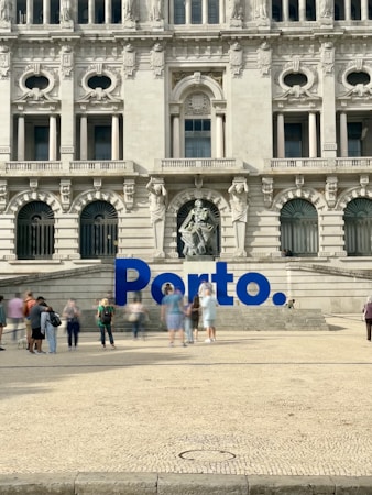 A large, ornate building with intricate architectural details and multiple arched windows serves as the backdrop. In front of the building, there is a prominent blue sign spelling 'Porto'. Several people are walking and standing around the area, creating a lively atmosphere.