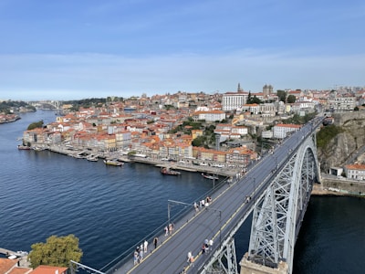 A bustling urban landscape featuring a large, arching iron bridge spanning a wide river. The bridge carries pedestrians and seems to connect two parts of a vibrant city. On one side of the bridge, a densely packed collection of buildings with red-tiled roofs is visible. The river flows smoothly under the bridge, reflecting the sky and cityscape.