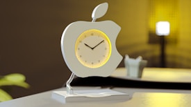A modern clock design shaped like an apple, featuring a silver frame with a leaf accent on top. The clock face is circular with black hour markers and hands, set against a backdrop of soft, warm lighting. The clock stands on a sleek metallic base, placed on a glossy surface that reflects the ambient light. In the background, there are soft blurred elements, including a light source and a plant, enhancing the contemporary aesthetic.