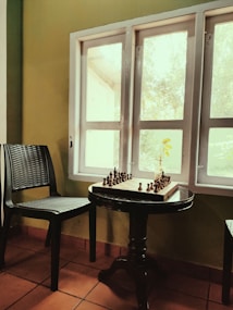 A cozy corner featuring a chessboard set on a round wooden table near a sunlit window. A single chair with a woven texture is positioned beside the table, and a small plant with yellow leaves is on the windowsill. The room has terracotta floor tiles and walls in a warm green tone.